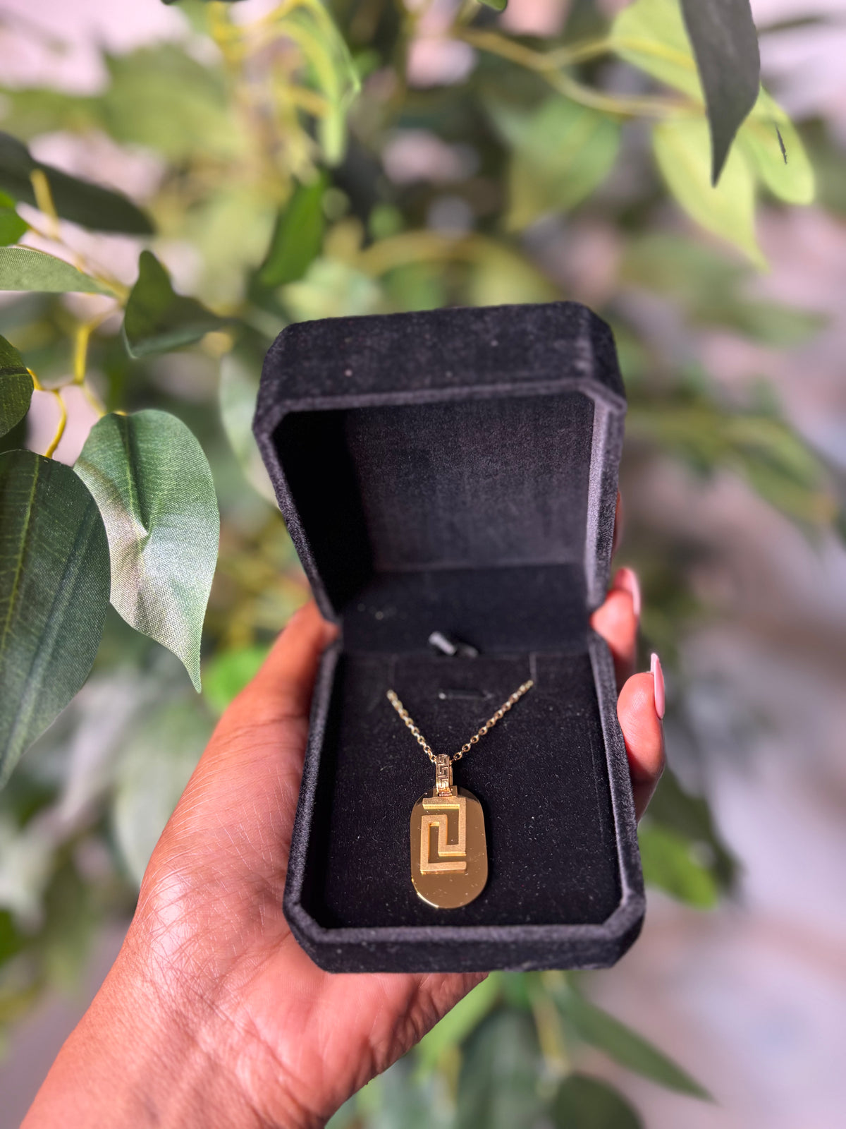 A gold pendant necklace with a geometric engraved design is displayed inside an open black velvet jewelry box, held by a hand against a blurred background of green leaves.